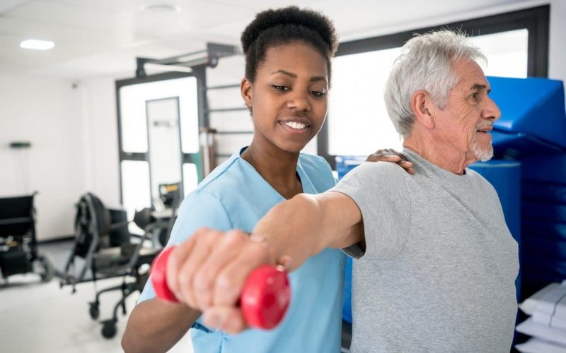 African american occupational therapist helping a senior patient with his shoulder workout at the clinic, both looking happy and smiling